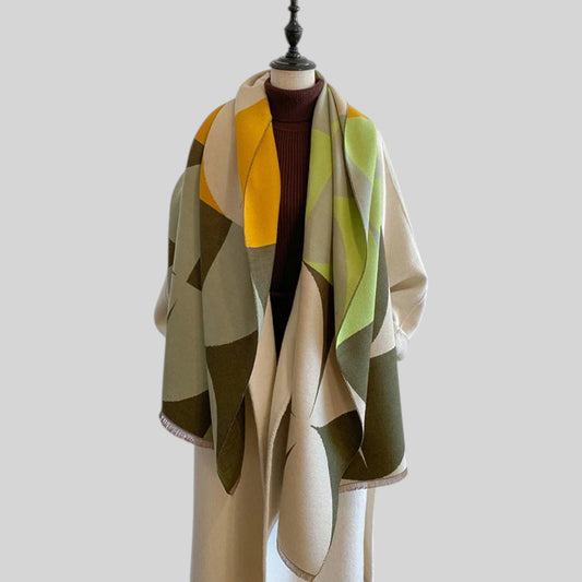 A large geometric patterned scarf draped over a cream-colored mannequin torso, styled with a dark brown ribbed turtleneck underneath. The scarf features large color-blocked shapes in cream, olive green, mustard yellow, and light green, with a small fringe detail along the edge, set against a solid light gray background.