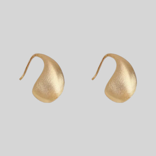 A pair of organic-shaped gold earrings with a textured, matte finish and curved hooks, shown on a solid light gray background.