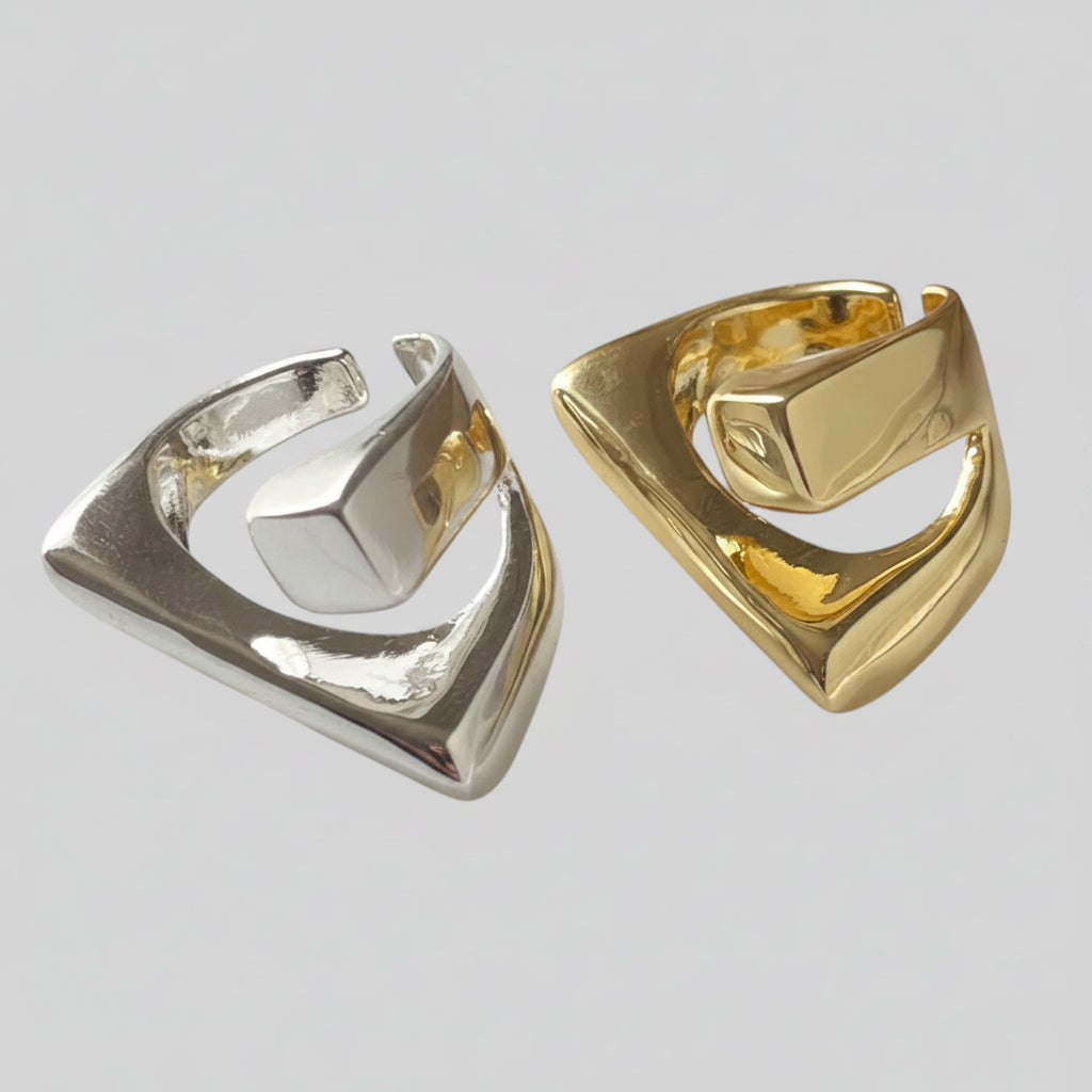Two abstract geometric hoop earrings shown side by side on a light gray background. One earring is polished silver-toned metal, and the other is polished gold-toned metal.
