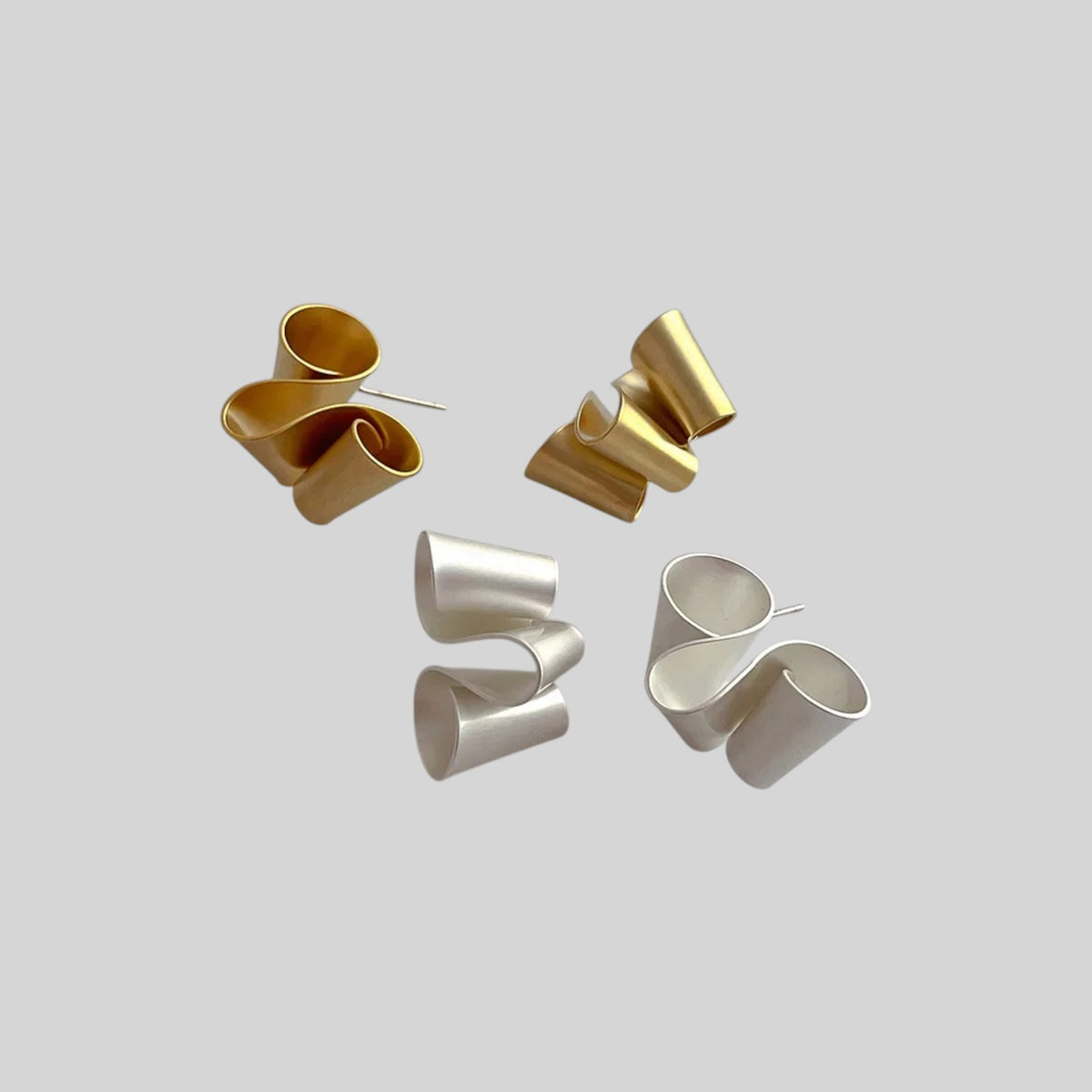 Four sculptural, curved metal rings in polished gold and silver finishes, arranged on a solid light gray background.