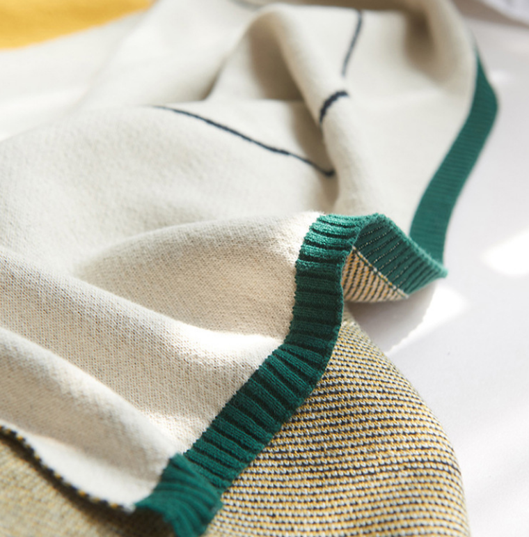 Close-up of a knitted textile with a cream and beige body, dark green ribbed trim, and thin dark line details, photographed on a light background.