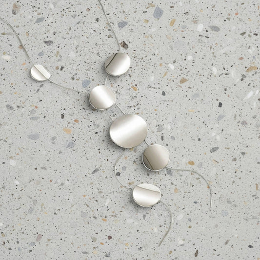 A minimalist silver disc pendant necklace with five graduated oval discs on a thin chain, laid out on a speckled gray terrazzo surface.