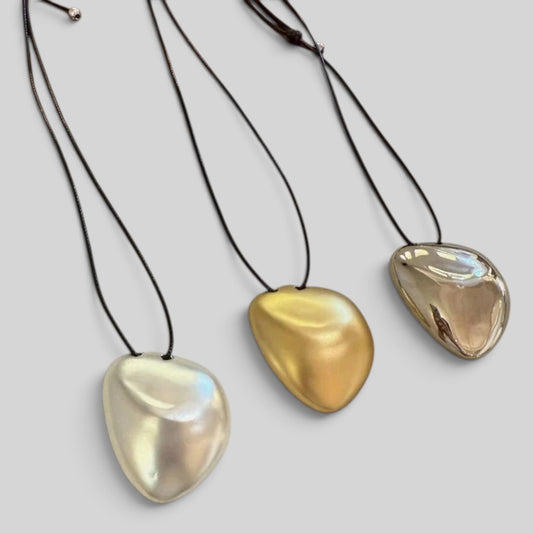 Three smooth teardrop-shaped pendant necklaces on black cords, shown against a light gray background. The pendants are pearlescent white with iridescent blue highlights, polished gold, and metallic rose gold.