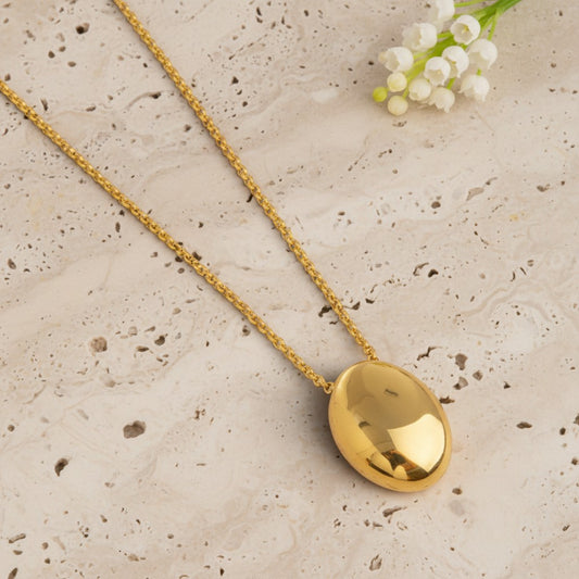 A gold chain necklace with a polished oval pendant, displayed diagonally on a textured beige stone surface with small white flowers in the corner.