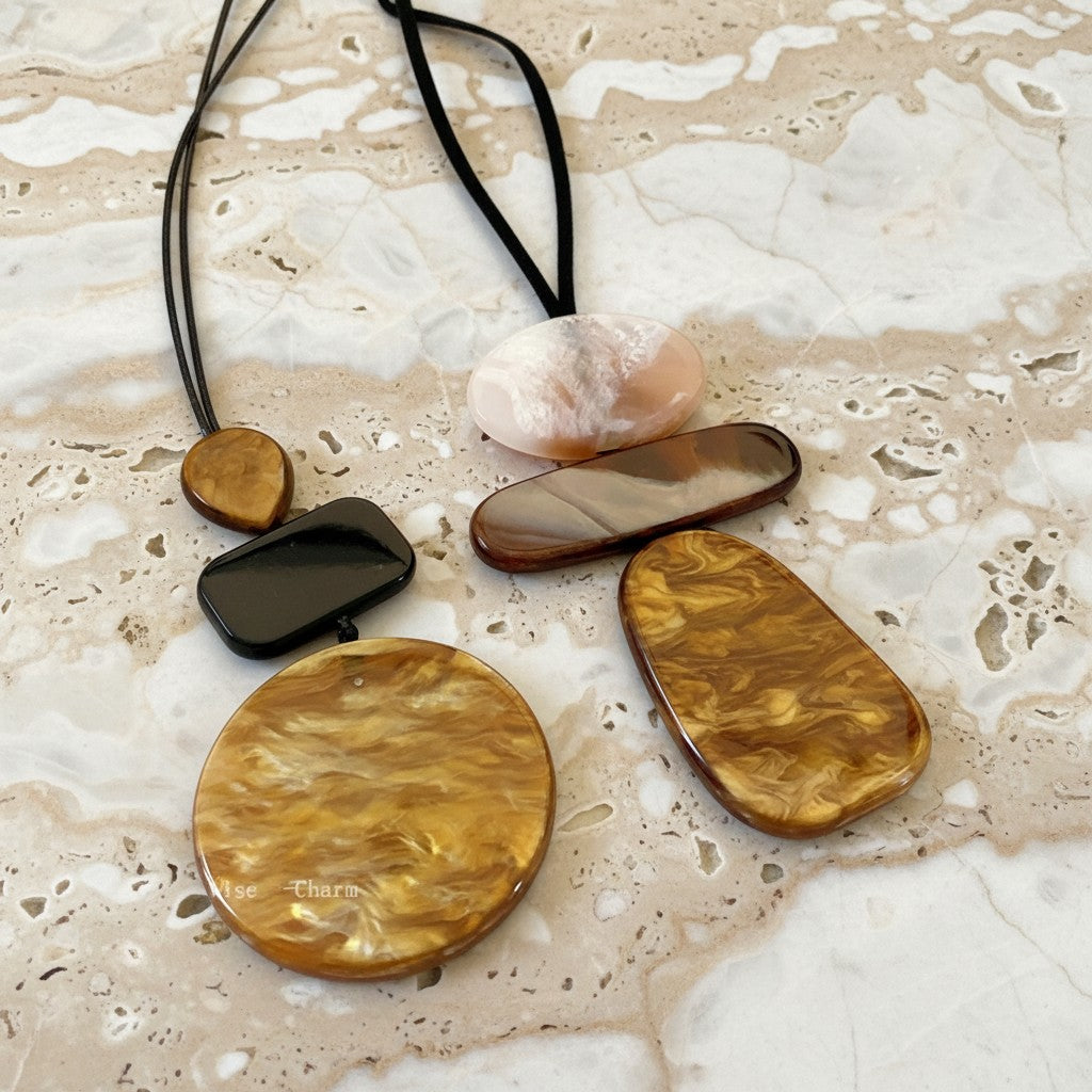 A necklace laid flat on a light beige and white marbled surface, featuring multiple polished stone-like pendants in amber, brown, black, and pale pink tones attached to a black cord.