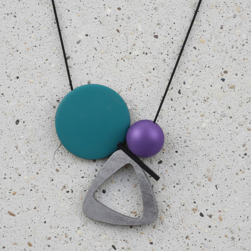 A modern geometric pendant necklace featuring a matte teal disc, a metallic purple sphere, and a textured gray triangular frame on a black cord, displayed on a speckled concrete surface.