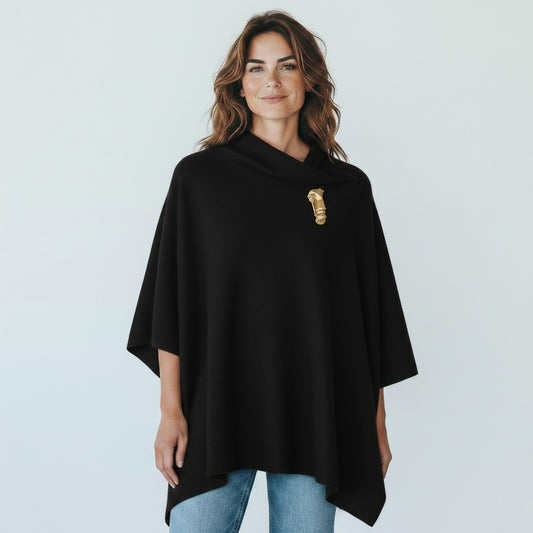 A woman wearing a black draped poncho-style top with a gold decorative accent on the chest, paired with blue jeans, against a light gray studio background.
