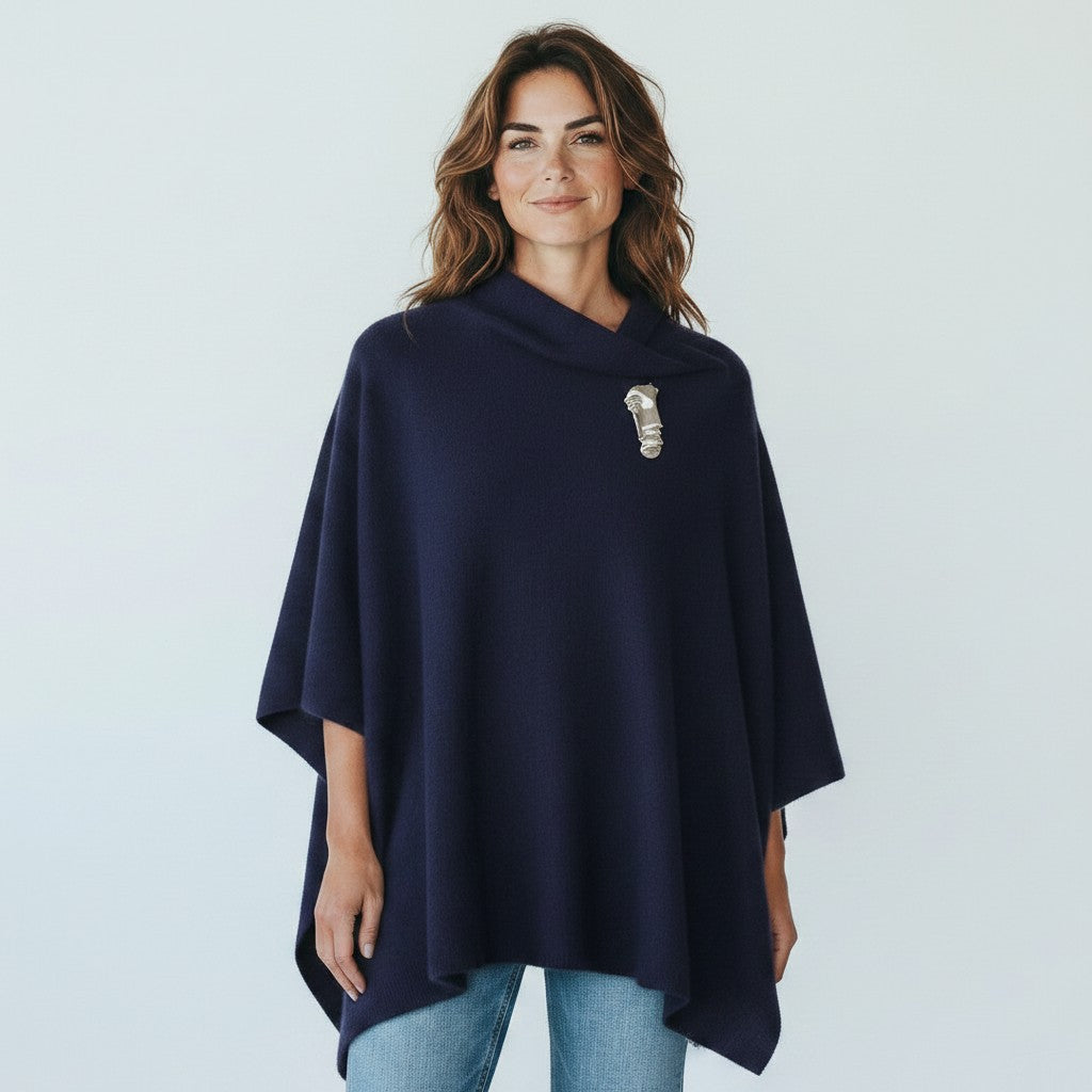 A woman wearing a navy blue cowl-neck poncho with a silver-toned pendant accessory at the neckline, paired with light blue jeans, against a light gray studio background.