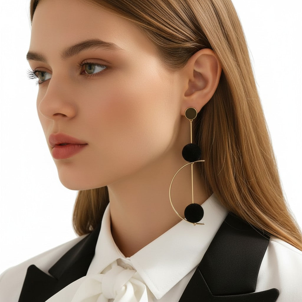 A close-up of a woman wearing a pair of long, statement dangle earrings. The earrings feature a gold-tone metal frame with three stacked black pom-pom spheres.