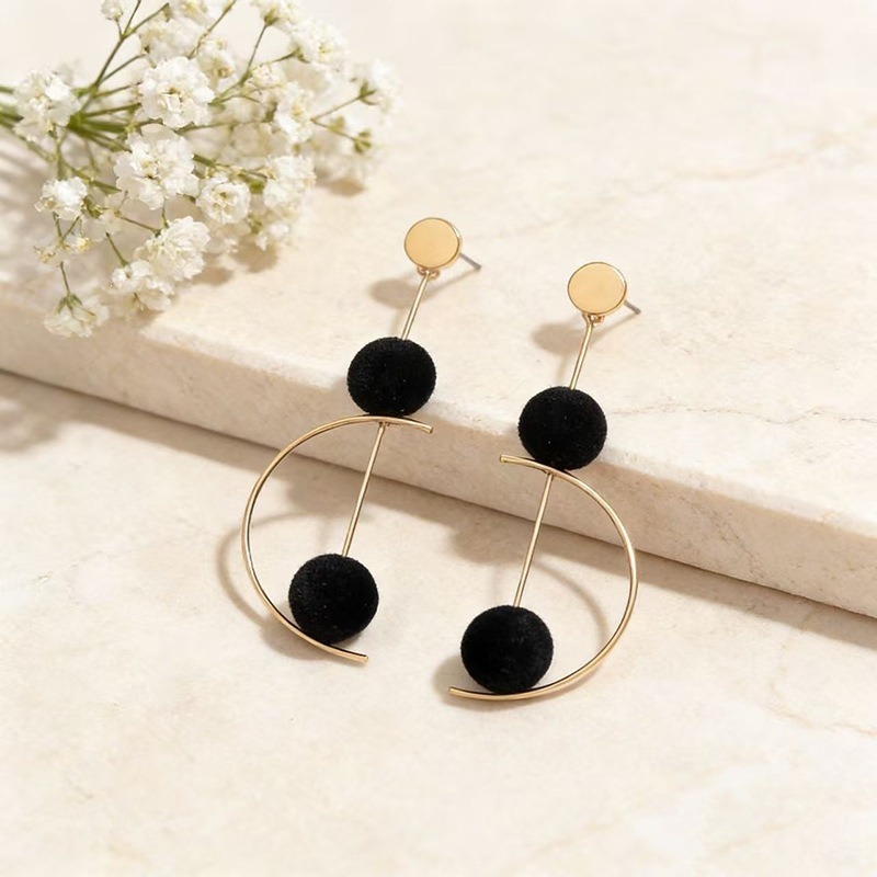 A pair of gold-tone geometric dangle earrings with black velvet spheres, displayed on a light marble surface next to small white flowers.