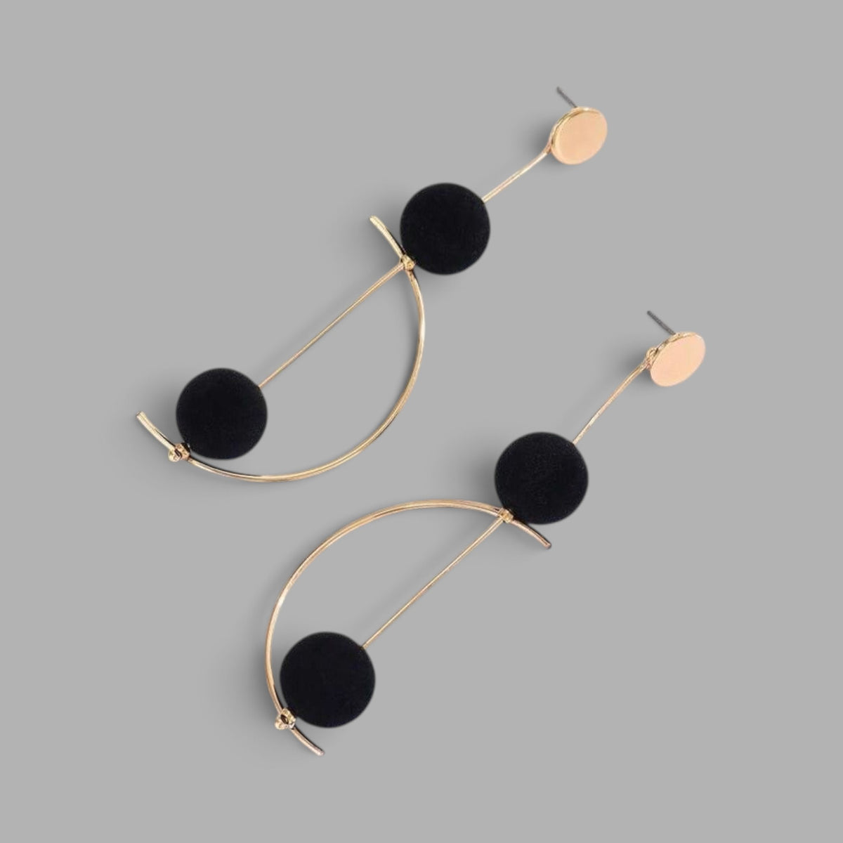 A pair of gold-tone geometric dangle earrings with black pom-pom spheres, displayed on a solid gray background.
