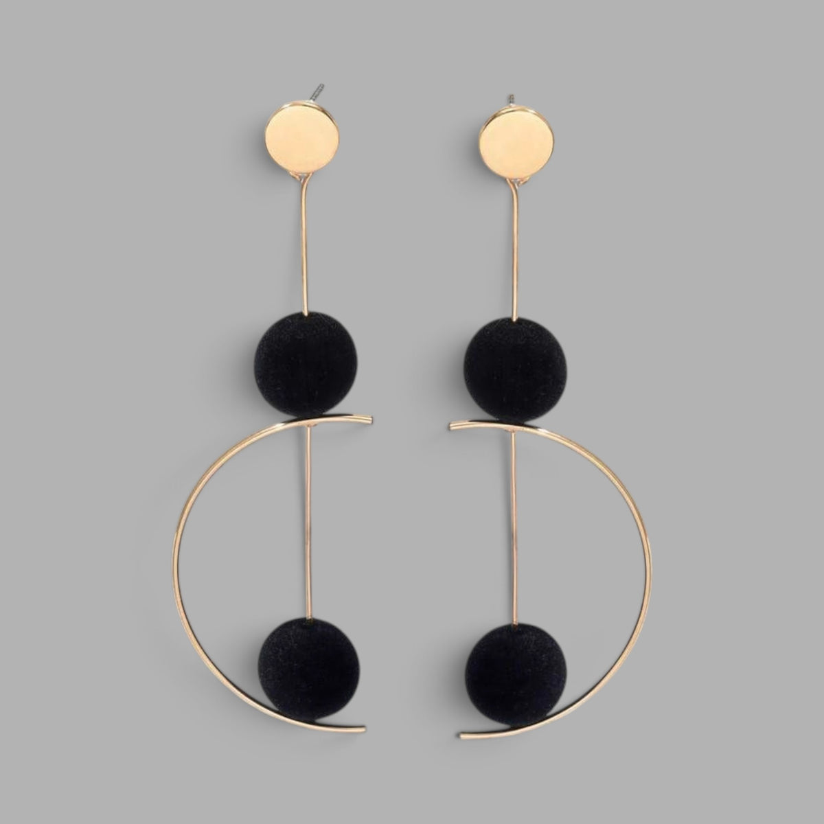 A pair of gold and black statement dangle earrings on a solid gray background. Each earring features a flat gold disc stud, a vertical gold bar, a black pom-pom ball, and a larger open-ended gold hoop with a second black pom-pom ball at the bottom.