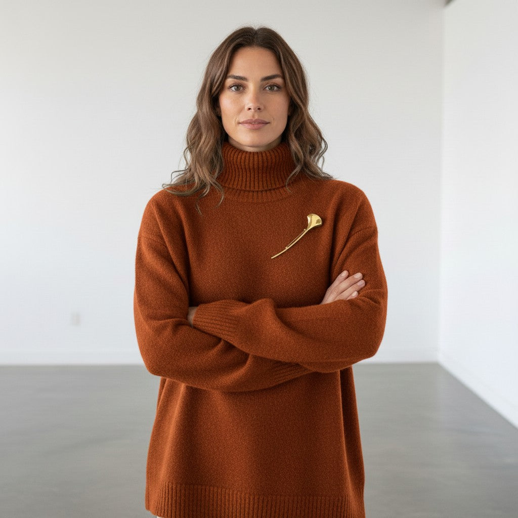 A woman with wavy brown hair stands with arms crossed, wearing a rust-orange turtleneck sweater with a small gold flower-shaped brooch on the chest, set against a minimalist white room with gray concrete floors.