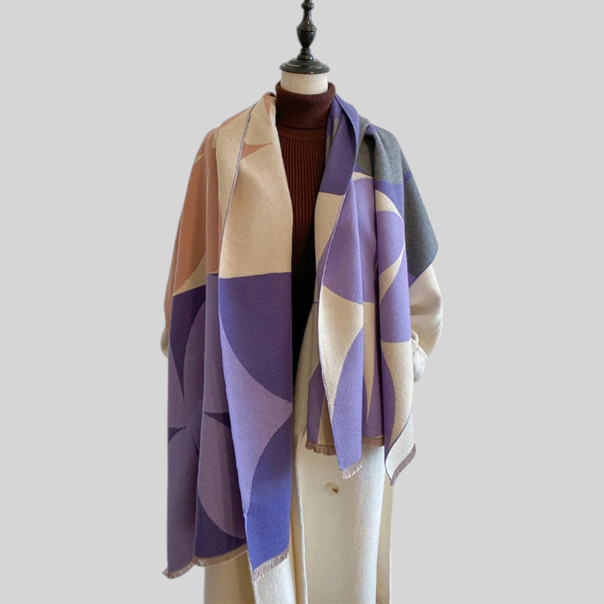 A multicolor geometric patterned knit scarf draped over a cream-colored coat and a dark brown ribbed turtleneck sweater, displayed on a mannequin torso against a solid light gray background.