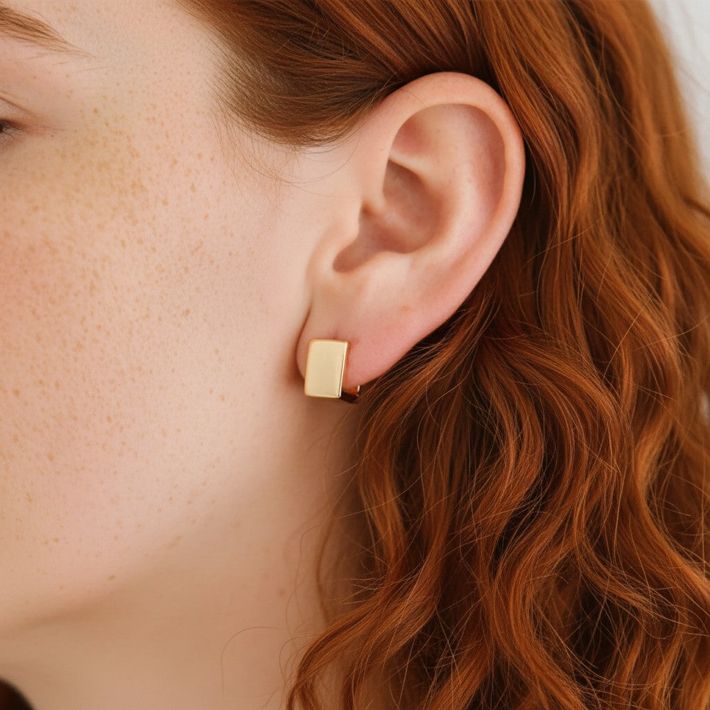 A close-up of a person's ear wearing a small rectangular gold stud earring with a tiny clear gemstone, against a soft light background.