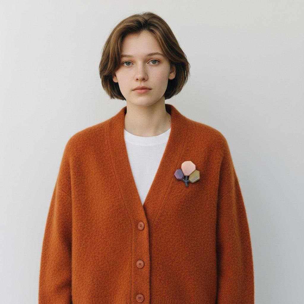 A young woman with short brown hair wearing a fuzzy rust-orange cardigan over a white t-shirt, featuring a small multi-colored flower pin on the chest, set against a plain light gray background.