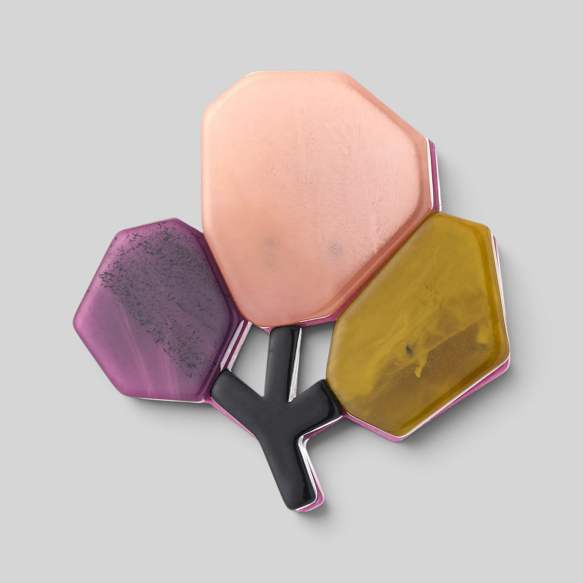 A three-sided geometric pin brooch with a central black Y-shaped metal stem and three faceted, translucent top plates in peach, purple, and mustard yellow, set against a solid gray background.