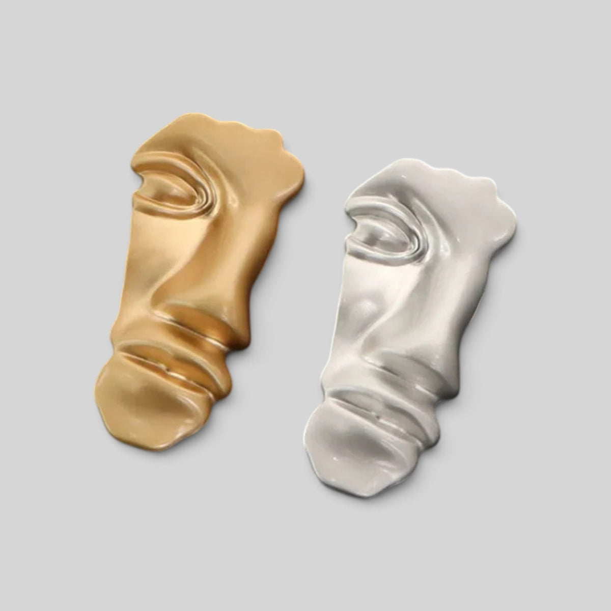 Two sculptural face-shaped accessories, one in metallic gold and one in metallic silver, displayed side-by-side on a solid light gray background.