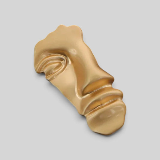 A gold-colored, abstract sculptural object with a glossy finish, photographed on a solid light gray background.