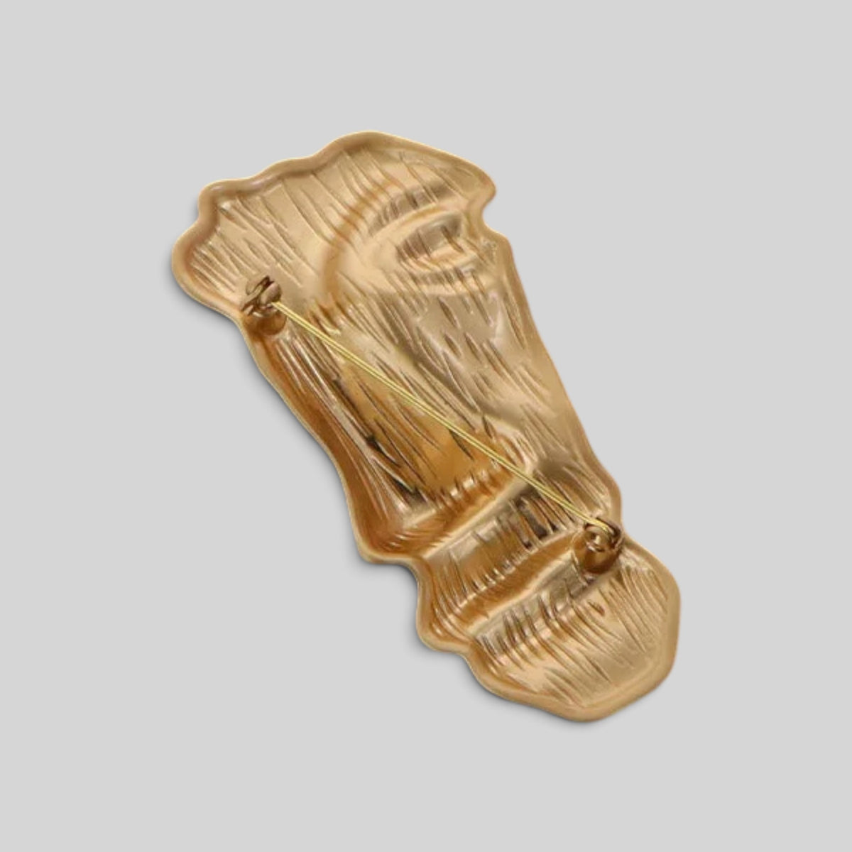 A gold-toned textured resin brooch shaped like a stylized face, shown from an angled perspective against a solid light gray background.