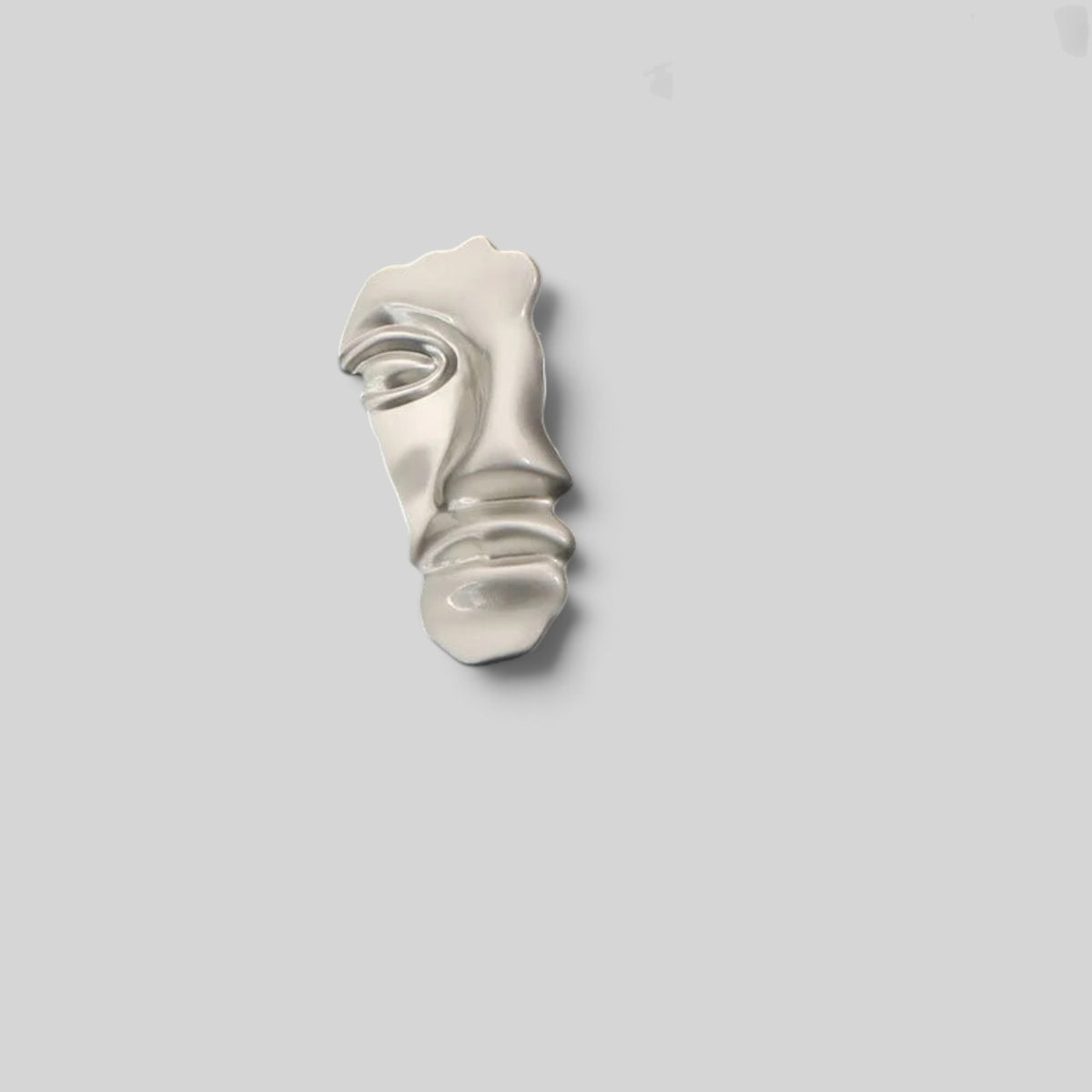 A small sculptural metallic object with an abstract face-like form, shown on a plain light gray background.