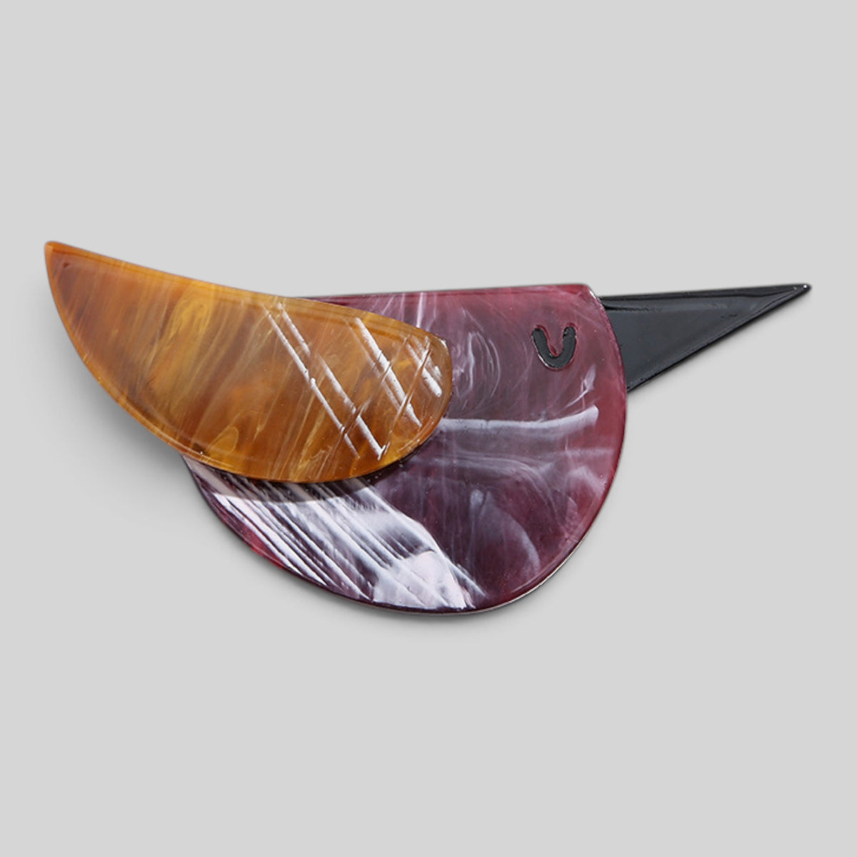 A sculptural, bird-shaped resin pin or brooch featuring a translucent amber-brown wing, a marbled burgundy and white body, and a black beak, shown on a neutral gray background.