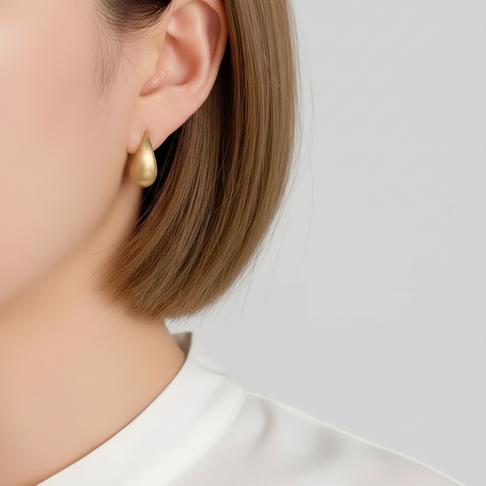 A close-up of a model's ear wearing a small, teardrop-shaped gold earring, with her hair and white top visible against a light gray background.