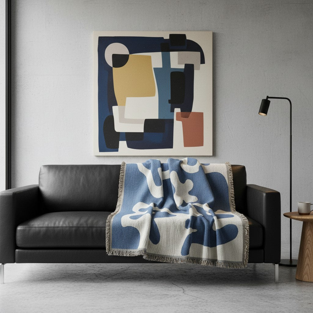A modern living room scene featuring a black leather sofa with a blue and white patterned throw blanket, beneath a large abstract geometric wall art print on a light gray wall.