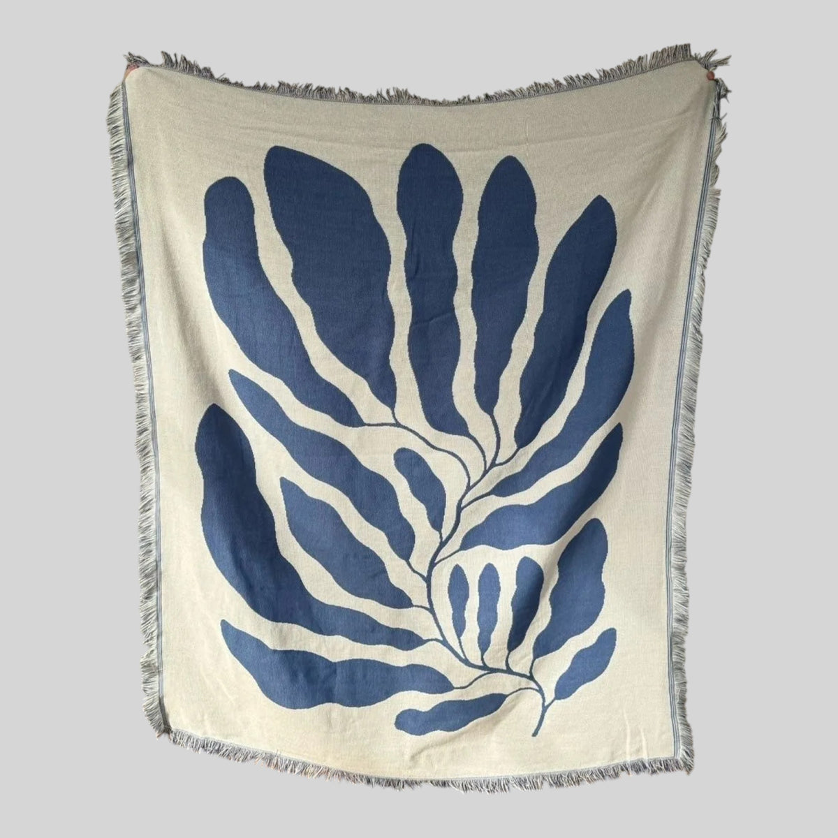 A square cream-colored woven throw blanket with fringed edges, featuring a large navy blue botanical leaf pattern printed on the front, shown against a neutral gray background.