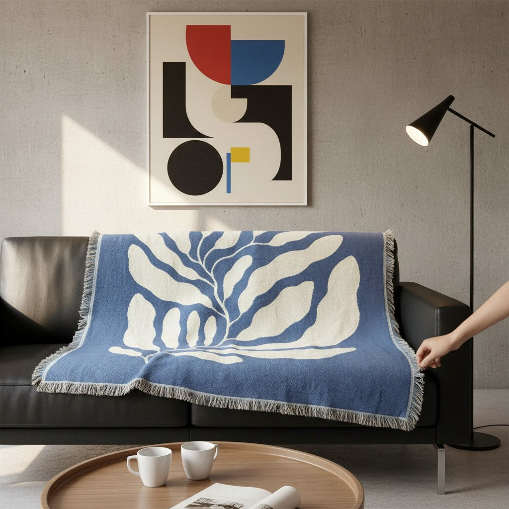 A blue and white woven throw blanket with a leaf pattern and fringe is draped over a black leather sofa in a modern living room setting. A person's arm is visible holding the corner of the blanket.