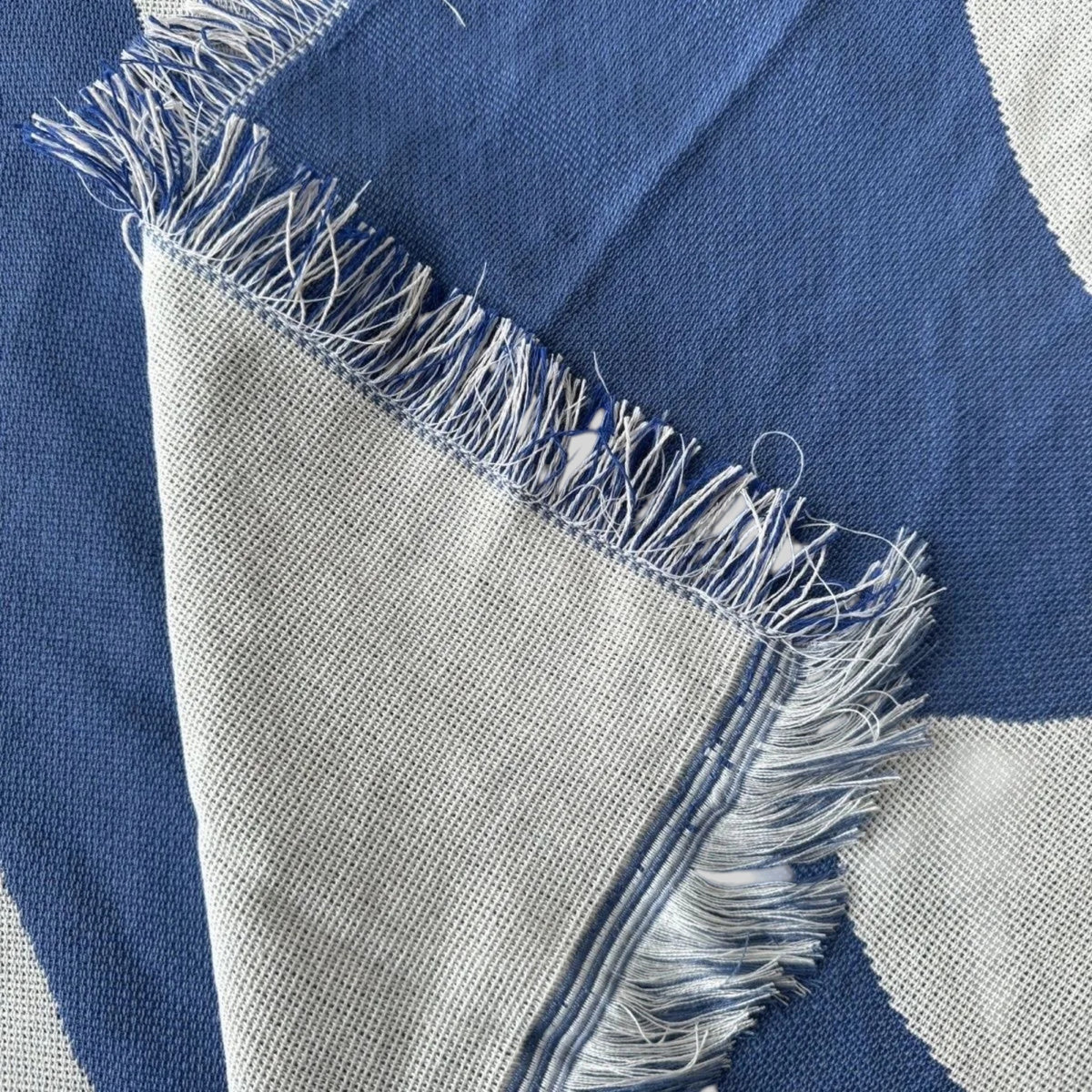 A close-up of a woven textile with a blue and off-white pattern, featuring frayed fringe along the folded edge.