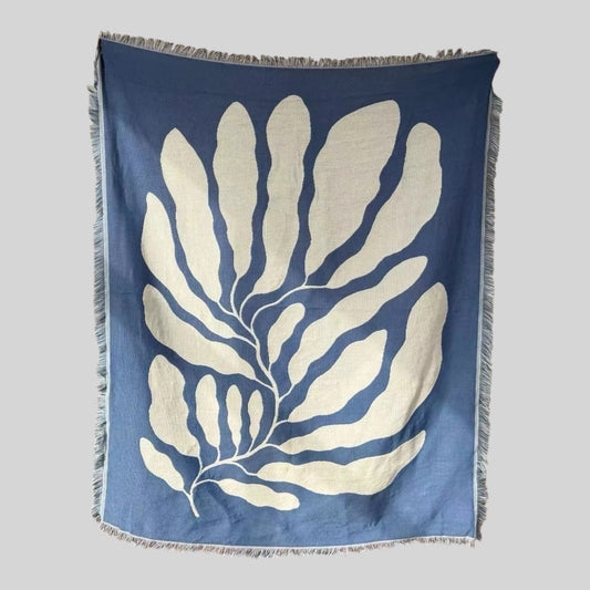 A square woven textile with fringed edges, featuring a large off-white botanical leaf pattern on a muted blue background, shown against a plain light gray backdrop.