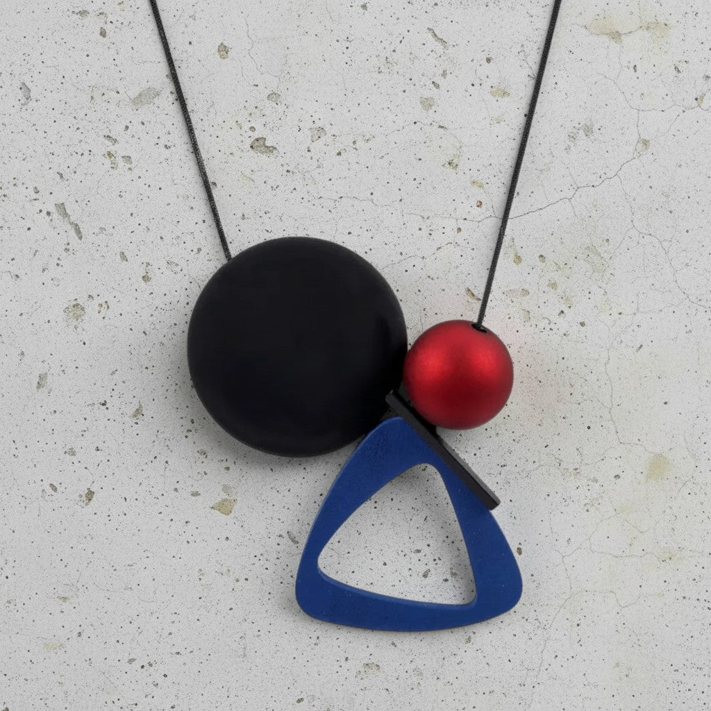 A modern geometric pendant necklace featuring a matte black oval, a metallic red sphere, and a blue triangular frame on a black cord, displayed on a textured light gray surface.