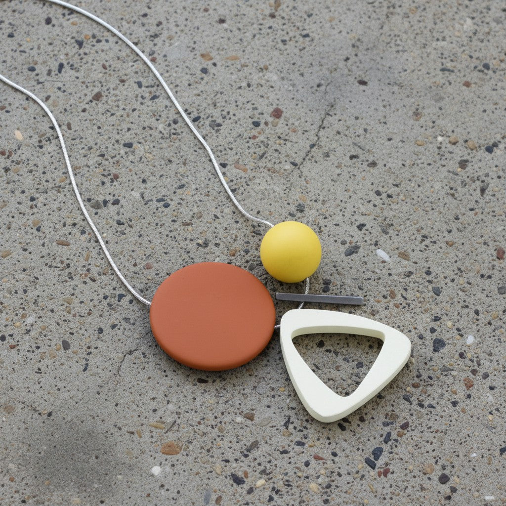 A geometric pendant necklace on a concrete surface, featuring a terracotta-colored flat circle, a small yellow sphere, a white triangular frame, and a thin silver wire chain.
