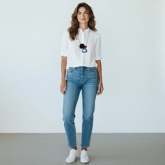 A woman stands against a plain white wall wearing a white short-sleeve button-up shirt, light-wash blue jeans, and white sneakers. She is also wearing a long black pendant necklace with colorful geometric charms.