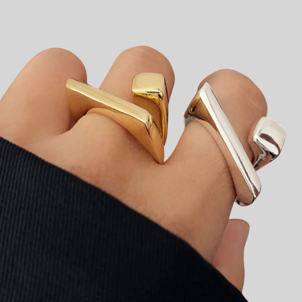 A close-up of a hand wearing two chunky geometric shift rings, one in polished yellow gold and the other in polished silver, against a neutral light gray background.