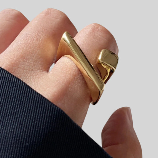A close-up of a hand wearing a chunky, modern gold ring with a geometric, angular design and a polished finish, set against a neutral light gray background.