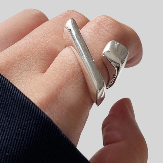 A close-up of a hand wearing a chunky silver ring with a diagonal bar and a separate sculptural side piece, shown against a light gray background.