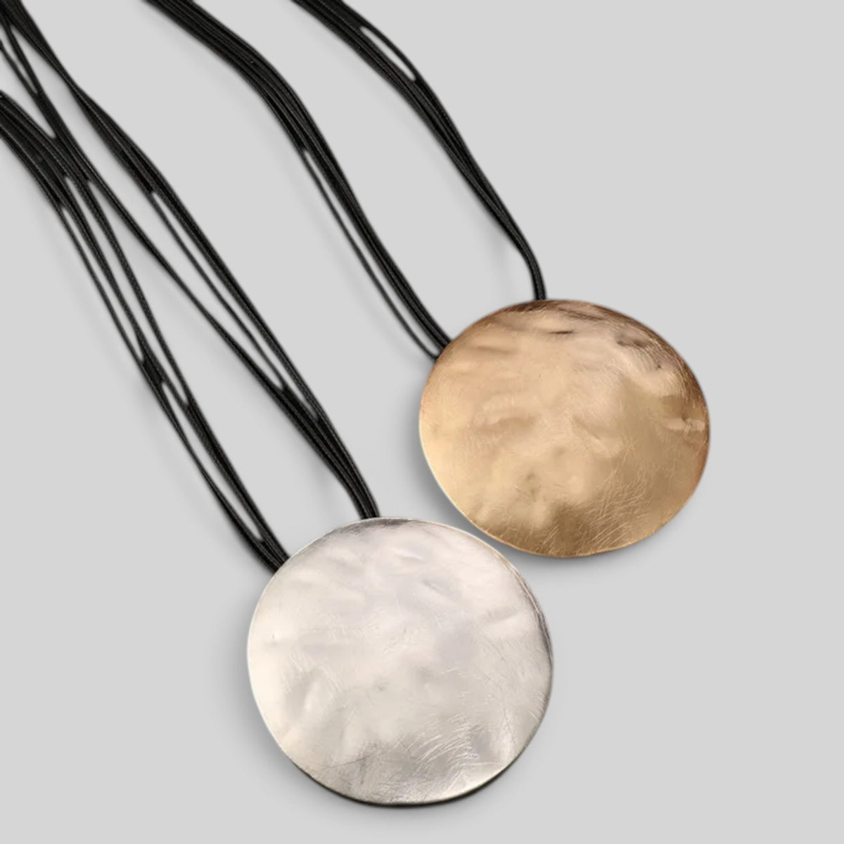 Two round hammered metal pendant necklaces on black cords, one in silver and one in gold, displayed on a light gray background.