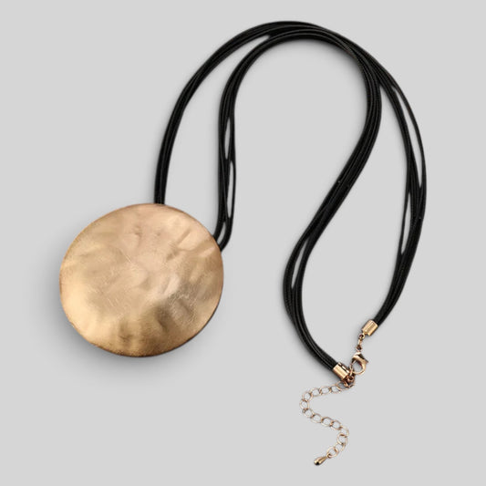 A modern statement necklace featuring a large, hammered round gold disc pendant on a multi-strand black cord, with a gold clasp and extender chain, shown on a neutral gray background.