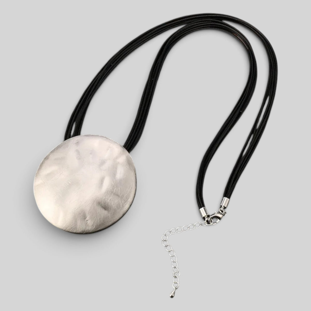 A silver pendant necklace with a textured, hammered circular disc on a black cord, featuring a small silver chain extender, set against a solid gray background.