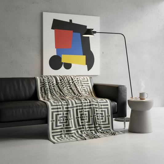 A modern living room scene with a black leather sofa draped in a black-and-white geometric patterned throw blanket, a black floor lamp, a small gray side table with a mug, and a large abstract wall art print above the sofa.