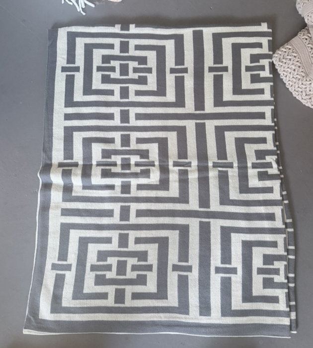 A square woven textile with a bold gray and off-white geometric maze-like pattern, laid flat on a dark gray surface.