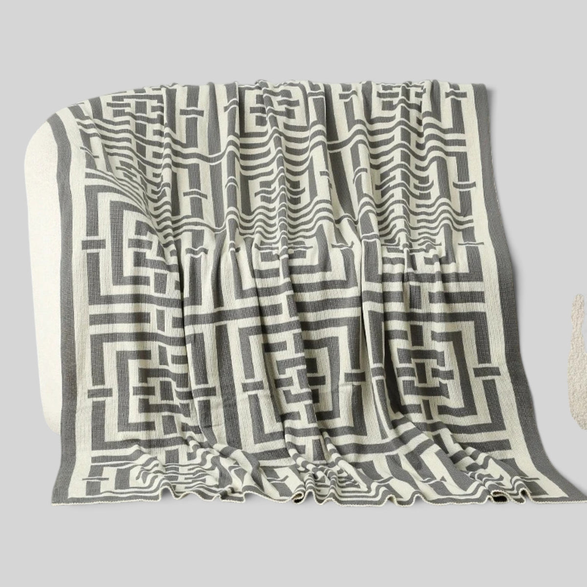 A woven throw blanket with a geometric maze-like pattern in dark gray and off-white, draped to show its texture and pattern against a solid gray background.