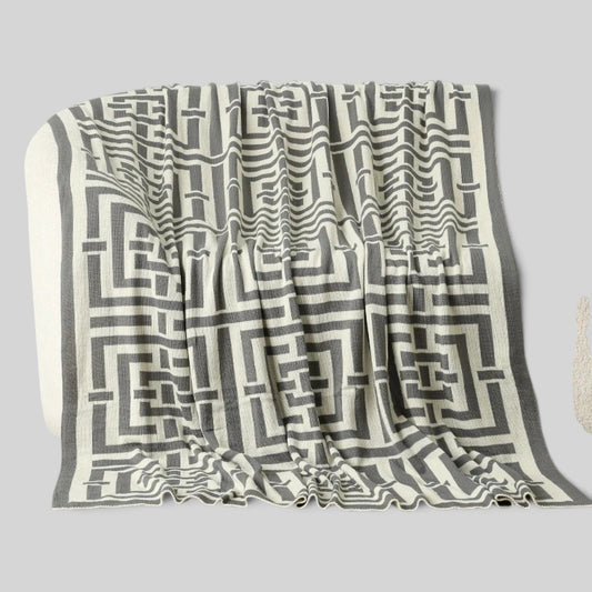 A woven throw blanket with a geometric maze-like pattern in dark gray and off-white, draped to show its texture and pattern against a solid gray background.