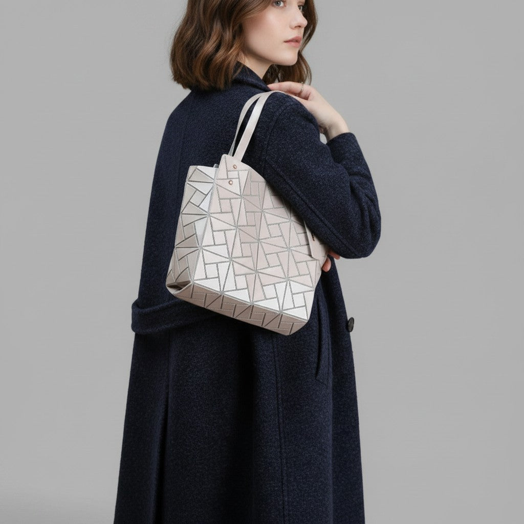 A woman in a dark navy coat carries a light beige geometric tote bag with a silver geometric pattern, photographed against a solid gray studio background.