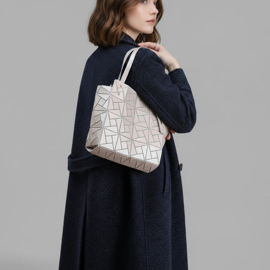 A woman in a dark navy coat carries a light beige geometric tote bag with a silver geometric pattern, photographed against a solid gray studio background.