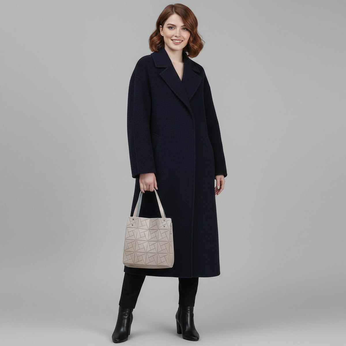 A woman stands against a light gray studio background wearing a long navy coat, black pants, black ankle boots, and holding a structured light beige handbag with geometric embossed detailing.