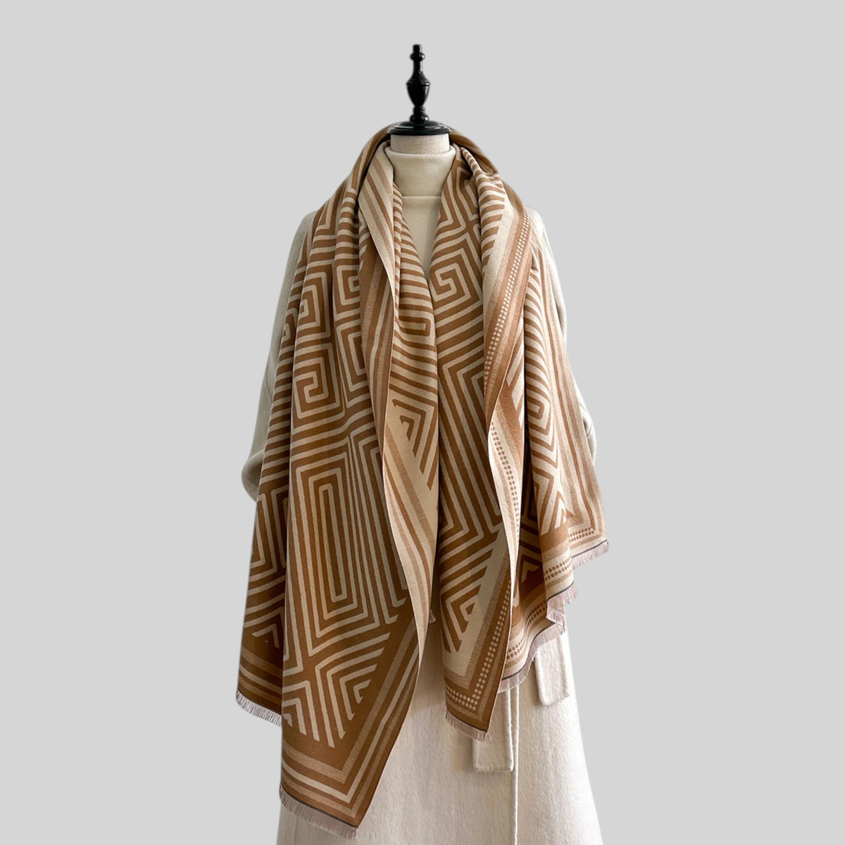 A large geometric patterned scarf in shades of brown and beige, draped over a cream-colored dress form against a solid light gray background.