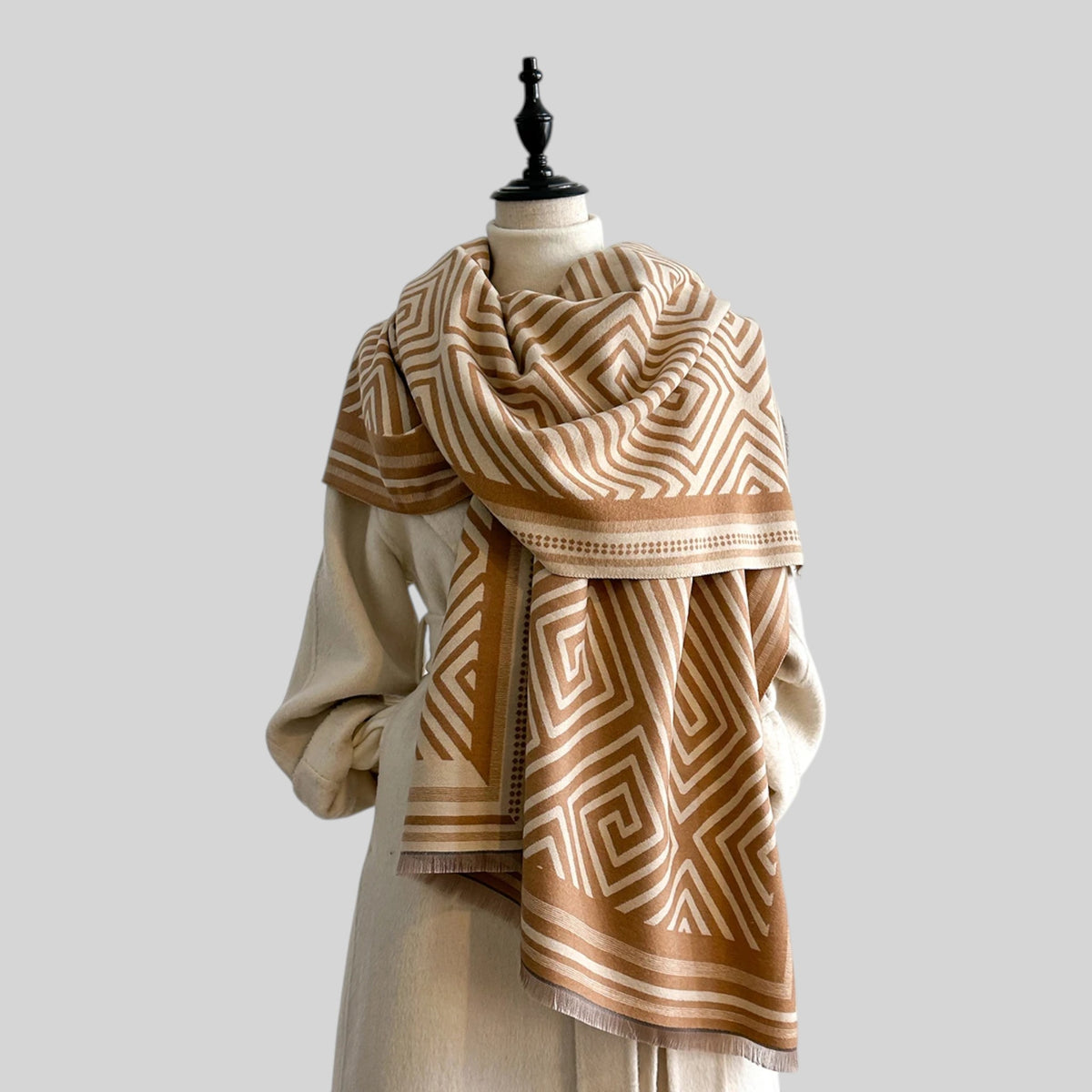 A beige and brown geometric patterned scarf draped on a cream-colored mannequin torso against a solid light gray background.